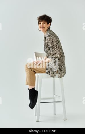Eine stilvolle Frau mittleren Alters mit kurzen Haaren sitzt in einem Studio auf einem Hocker. Stockfoto