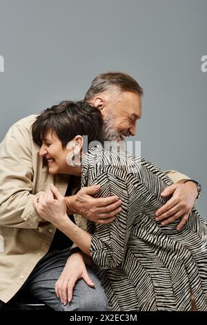 Ein Mann mittleren Alters umarmt liebevoll eine Frau von hinten auf einem Stuhl in einem stilvollen Studio-Setting. Stockfoto