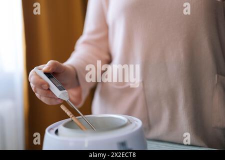 Nahaufnahme von nicht erkennbaren weiblichen Handwerkstemperaturmessungen von geschmolzenem Wachs. Prozess der Herstellung handgemachter, natürlich duftender Kerzen bei Stockfoto