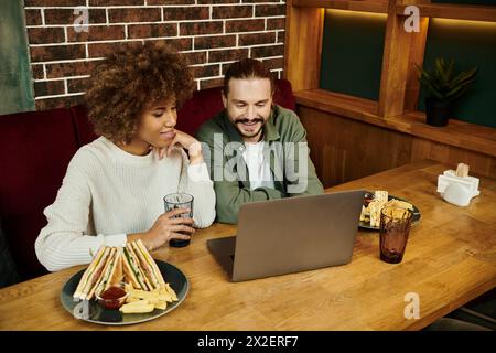 Eine Afroamerikanerin und ein Mann sitzen an einem Tisch in einem modernen Café, konzentriert auf den Laptop-Bildschirm vor ihnen. Stockfoto