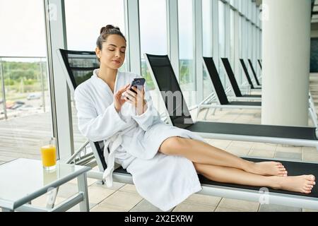 Eine junge brünette Frau im Bademantel entspannt sich in einem Wellnessbereich, während sie ein Handy benutzt. Stockfoto