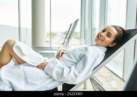 Eine junge, schöne Brünette in einem Bademantel sitzt friedlich in einem Liegestuhl drinnen in einem Spa mit Swimmingpool. Stockfoto