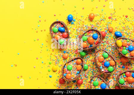 Schokoladeneier mit Süßigkeiten und zuckerfarbenen Streuseln auf gelbem Hintergrund, Blick von oben. Osterkomposition. Hälften Schokoladeneier auf dem Tisch. F Stockfoto