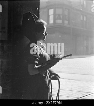 CECIL BEATON PHOTOGRAPHS: TYNESIDE SHIPYARDS, 1943 - Eine Studie einer jungen Schweißerin. Sie posiert für den Fotografen und blickt lächelnd weg, während sie sich gegen eine Säule lehnt. Im Hintergrund ist eine Straße mit Wohngebäuden zu sehen Stockfoto