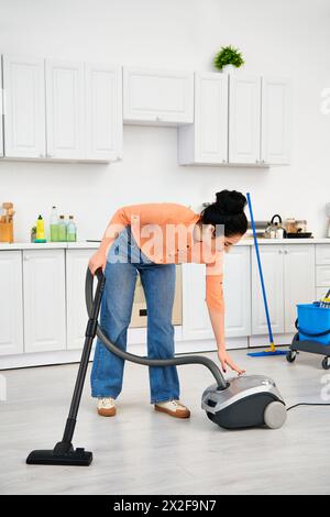 Eine stilvolle Frau in lässiger Kleidung reinigt leidenschaftlich ihren Küchenboden mit einem Staubsauger. Stockfoto