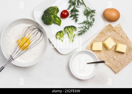 Zutaten für die Teigherstellung. Gebrochenes Ei mit Mehl in einer Schüssel. Auf die Schüssel rühren. Butter auf Papier. Brokkoli, Tomaten und Dill in Teller. Weißer Hintergrund. F Stockfoto
