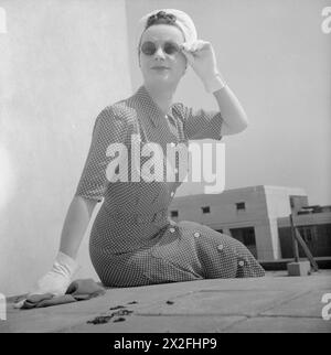 WIE Eine BRITISCHE FRAU SICH IN KRIEGSZEITEN KLEIDET: GEBRAUCHSKLEIDUNG IN GROSSBRITANNIEN, 1943 – Ein Model sitzt an einer Wand, als sie für die Kamera auf einem Dach in Bloomsbury posiert, um ihr rotrotes und weißes gepunktetes Utility-Hemdkleid mit Knopfleiste vorne zu zeigen. Dieses Kleid kostet 7 Coupons und 53/-. Sie trägt das Kleid mit einem weißen Turban (auch in D 14782, D 14786 und D 14798) und weißen Handschuhen. Sie trägt auch eine Sonnenbrille, ihre Handschuhe halten den Drahtarm der Brille Stockfoto