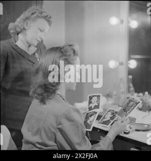 GEBURT Eines STARS: ALLTAG FÜR SCHAUSPIELERIN MURIEL PAVLOW, ENGLAND, UK, 1945 - Schauspielerin Muriel Pavlow (sitzend, nächstgelegene Kamera) und Friseurin Elsie Bass schauen sich Fotos verschiedener Frisuren an, die sie für Muriel in einer Ankleidekammer ausprobiert haben, wahrscheinlich in den Welwyn Garden Studios, Hertfordshire. Laut der Originalüberschrift schenkt die Filmfirma dieser wichtigen Rolle beim Aufbau eines Stars große Aufmerksamkeit. Ihre Entscheidung, die endgültig ist, stimmt nicht immer mit der Meinung des STAR überein." Stockfoto