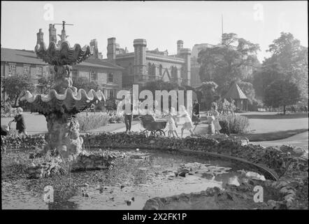 Ein BILD EINER SÜDLICHEN STADT: LIFE IN WARTIME READING, BERKSHIRE, ENGLAND, Großbritannien, 1945 - Kinder spielen, während sie um den Brunnen und den Lilienteich in einem sonnigen Forbury Gardens in Reading spazieren. Eines der Mädchen schiebt einen Kinderwagen, während sie läuft. Ebenfalls sichtbar ist ein Soldat, der einen Spaziergang in der Sonne macht. Der Originaltitel besagt, dass „unter den rosa und weißen Seerosen eine Menge Goldfische sind“ Stockfoto