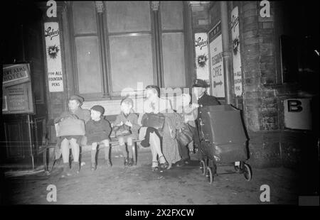 BRITISH RAILWAYS AT WAR - CHANGE AT CREWE: LIFE AT CREWE RAILWAY STATION, CHESHIRE, ENGLAND, UK, 1944 - in den frühen Morgenstunden sitzen ein Mitglied der Royal Navy und seine Familie auf einer Bank vor den Erfrischungsräumen am Bahnhof Crewe und warten auf einen Verbindungszug. Die Familie, zu der drei Kinder gehören, die auf der Bank sitzen und möglicherweise ein weiteres in einem Kinderwagen, ist aus Nordirland gereist Stockfoto