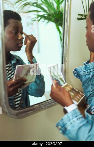 Junge kurzhaarige afrikanische junge Frau schminkt sich vor einem Spiegel und trägt in der Zwischensaison lässige Jeansjacke und Hemd Stockfoto
