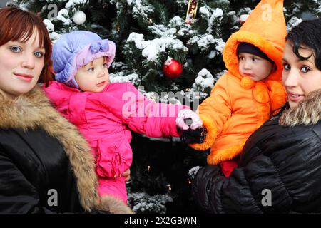 Zwei junge Frauen mit kleinen Töchtern stehen in der Nähe eines grünen Baumes mit Schnee; Kinder halten die Hände; konzentrieren Sie sich auf das Mädchen links Stockfoto