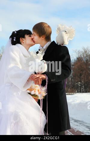 Schöne Braut und Bräutigam küssen und halten weiße Taube im Winter draußen; zweite weiße Taube sitzt auf der Schulter des Mannes Stockfoto