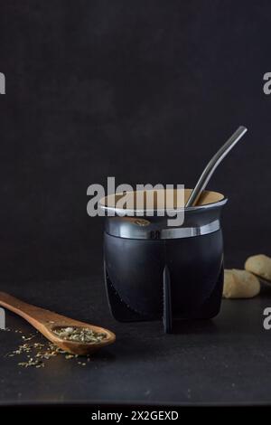 Yerba Mate Kraut und Kalabash Kürbis mit Stahlbombilla mit einem Holzlöffel und zwei Keksen auf schwarzem Hintergrund Stockfoto