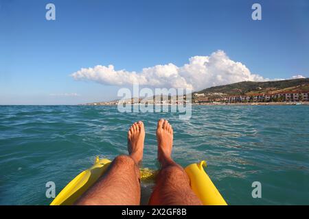 Herrenfüße auf gelber aufblasbarer Matratze, Meer, ferne Küste und Strand Stockfoto