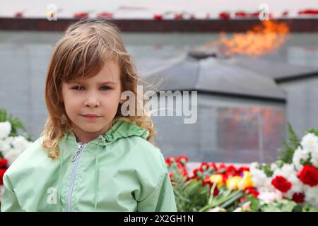 Das schöne kleine Mädchen steht in der Nähe der Gedenkstätte, ewige Flamme, Blumen Stockfoto