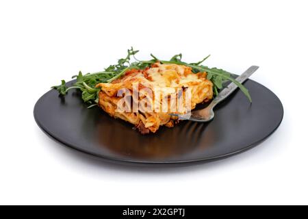 Quadratische Scheibe Lasagne auf einer schwarzen Keramikplatte mit Rucola-Salat, weicher Fokus aus der Nähe isoliert auf weiß Stockfoto