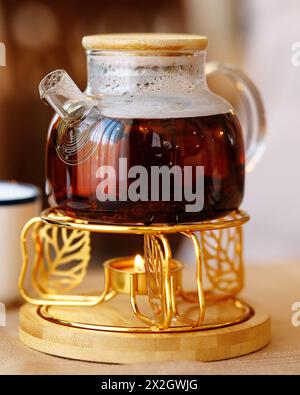 Teekanne aus Glas auf einem Stand, der von einer Kerze erhitzt wird. Schwarzer Tee in einer Teekanne aus Glas auf dem Tisch. Selektiver Fokus. Stockfoto