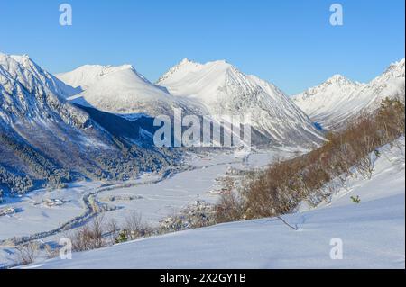 Ein malerischer Blick auf ein Dorf in einem Tal mit schneebedeckten Bergen unter einem klaren blauen Himmel. Stockfoto