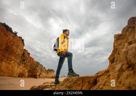Wanderer mit Rucksack, die Felsen am Strand hochlaufen, mit gelbem Mantel. Stockfoto