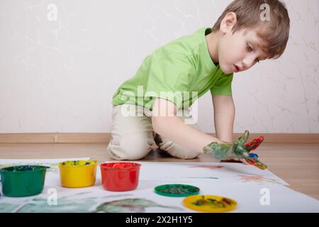 Der Junge im grünen T-Shirt zeichnet mit seinen Fingern Farbfarben auf das Blatt Papier Stockfoto