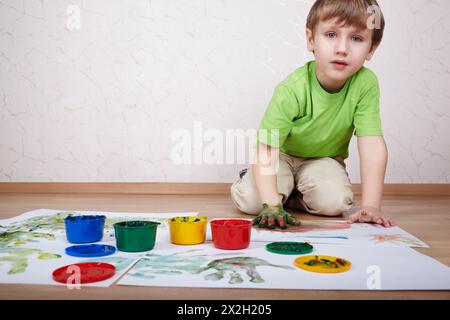 Junge im grünen T-Shirt zeichnet Farbfarben und macht Handabdrücke auf weißem Papier Stockfoto