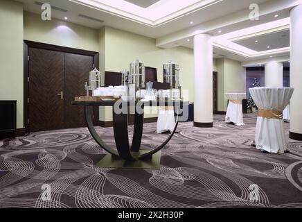 Großer Wasserkocher mit Geschirr auf der Stehparty zwischen den Tischen im geräumigen Saal bei der Konferenz Stockfoto