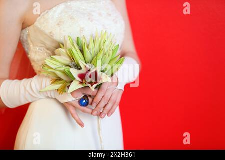 Die Hände der Braut in weißem Kleid halten einen Lilienstrauß auf rotem Hintergrund Stockfoto