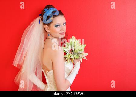 Lächelnde Braut mit blauem Make-up und Maske in der Frisur hält einen Strauß von Lilien auf rotem Hintergrund Stockfoto