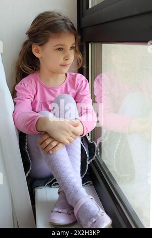 Das kleine hübsche Mädchen sitzt auf der Fensterbank und schaut aus dem Fenster Stockfoto