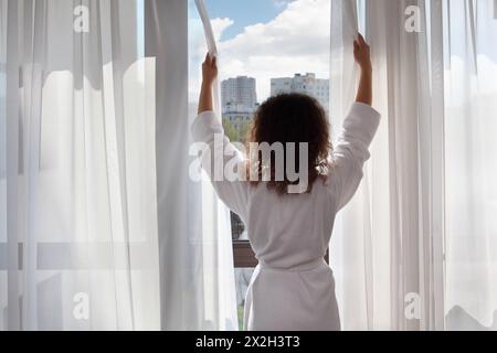 Frau in weißem Bademantel steht neben dem Fenster und öffnet die Vorhänge; Frau steht zurück zur Kamera Stockfoto