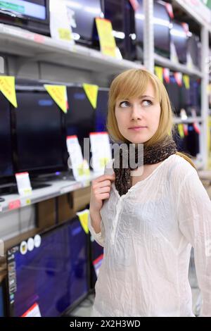 Blondes Mädchen mit Schal denkt darüber nach, im Supermarkt TV zu kaufen; geringe Schärfentiefe Stockfoto