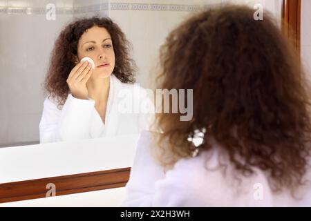 Lächelnde Frau in weißem Bademantel steht neben dem Spiegel und entfernt das Make-up Stockfoto