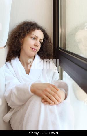 Eine wunderschöne Frau in weißen Bademantel sitzt auf der Fensterbank und blickt aus dem Fenster Stockfoto