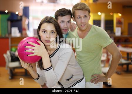 Die Frau bereitet sich darauf vor, beim Bowling einen rosa Ball zu werfen; zwei Männer schauen sich das Ziel an; konzentrieren sich auf die Frau; geringe Tiefe des Feldes Stockfoto