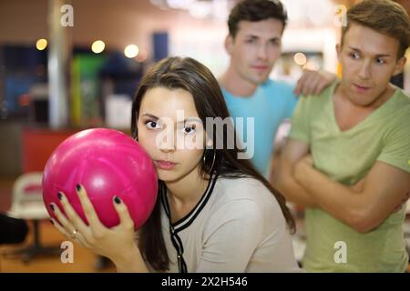 Die Frau bereitet sich darauf vor, beim Bowling einen Ball zu werfen; zwei Männer sehen sie an; konzentrieren sich auf die Frau; geringe Tiefe des Feldes Stockfoto