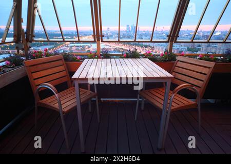 Holzstühle und Tisch auf der Terrasse im leeren Restaurant; Panoramablick auf Moskau am Abend Stockfoto