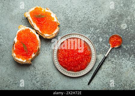 Kleine Metallplatte mit rotem Lachskaviar und zwei Kaviar-Toasts bieten Aussicht von oben auf grauem Betonhintergrund, festliche Luxus-Delikatesse und Vorspeise Stockfoto
