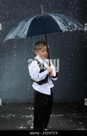 Junge in weißem Hemd gekleidet, steht unter Regenschirm im Regen und lächelt Stockfoto