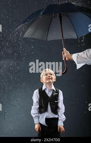 Lächelnder Junge in weißem Hemd und schwarzer Weste, der im Regen unter einem Regenschirm steht; die Hand des Mannes hält den Regenschirm Stockfoto