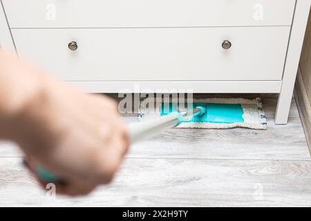 Die Hausfrau wischt den Boden unter dem Schrank. Nassreinigung des Fußbodens im Zimmer. Reinigung von Staub unter Schränken. Stockfoto