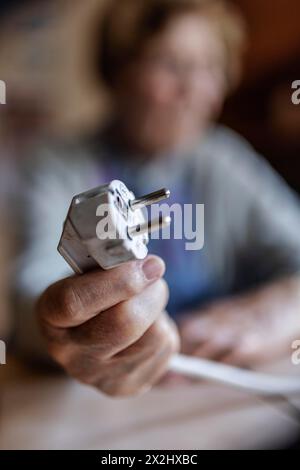 Seniorin, die zu Hause ein Netzkabel mit Stecker in der Hand hält, symbolisiert Energiekosten und Armut, Köln, Nordrhein-Westfalen, Deutschland Stockfoto