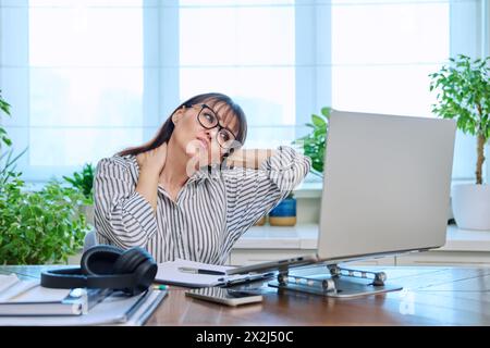 Müde Frau mittleren Alters sitzt an ihrem Arbeitsplatz, Schreibtisch mit Computer Stockfoto