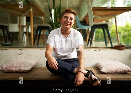 Lächelnder Gen Z Transgender-Büroangestellter in eleganter Freizeitkleidung sitzt auf einer Bank, in einer modernen Arbeitsumgebung. Tätowierte geschlechtsfließende Personen-Displays Stockfoto