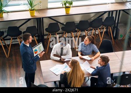 Die arabische Führungskraft präsentiert den Kollegen Verkaufsdaten auf einem Tablet in einem modernen Besprechungsraum. Multiethnische Teams setzen sich mit bunten Diagrammen und Diagrammen auseinander. Kreativ Stockfoto