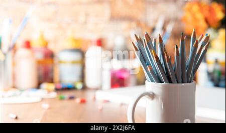 Weißer Becher mit farbigen Bleistiften im Vordergrund auf einem Holztisch mit Kunstzubehör im Hintergrund unscharf Stockfoto