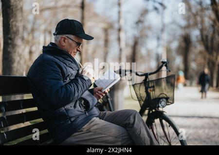 Ein älterer Herr entspannt sich auf einer Parkbank in der Ruhe eines sonnigen Tages, vertieft in das Lesen eines Buches, während sein Fahrrad neben ihm geparkt ist. Stockfoto