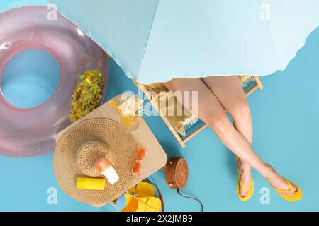 Junge Frau, die im Liegestuhl mit einem Glas Cocktail und Strandaccessoires auf blauem Hintergrund sitzt Stockfoto