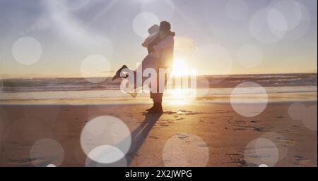 Bild von Lichtflecken über dem glücklichen afroamerikanischen Bräutigam, das Braut am Sonnenuntergang Strand bei Hochzeit hebt Stockfoto
