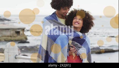 Bild von Lichtflecken über zweirassigen Paaren, bedeckt mit Decken am Strand Stockfoto